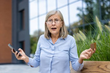 Kredi kartı ve telefon tutan endişeli bir son sınıf öğrencisi, dışarıda ofis binası yakınlarında yaptığı işlemlerle ilgili endişelerini dile getiriyor..