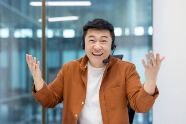 Asyalı bir adam, kulaklık takıyor, gülümsüyor ve ofis ortamında el kol hareketi yapıyor. Müşteri hizmetlerinde ya da çağrı merkezinde çalışıyor gibi görünüyor..
