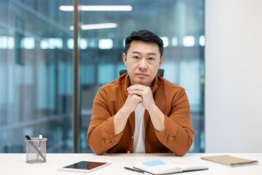 Odaklanmış Asyalı bir adam elleri bağlı bir masada oturuyor, muhtemelen modern bir ofis ortamında karar vermeyi düşünüyor..