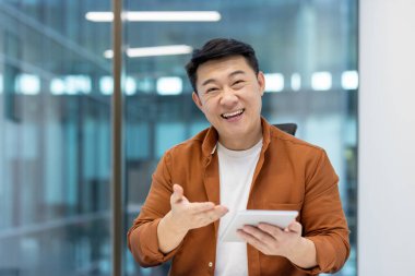 Parlak bir ofiste, elinde tabletle elini sallayarak gülümseyen mutlu Asyalı bir adam..