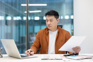 Asyalı bir işadamı dizüstü bilgisayar üzerinde çalışırken belgeleri inceliyor. O, masasında oturuyor..