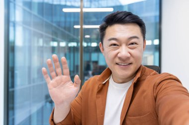 Sıcak bir gülümseme ile Asyalı bir adam kameraya el sallıyor, görünen o ki arka planda cam pencereler olan bir ofiste..