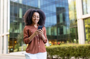 Gözlüklü, gülümseyen bir Afrikalı Amerikalı kadın, bir iş binasının yakınındaki bir şehir merkezinde akıllı telefonunu kontrol ediyor..