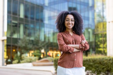 Gülümseyen siyah bir kadın modern bir binanın önünde duruyor, kollarını kavuşturmuş, güne hazır..