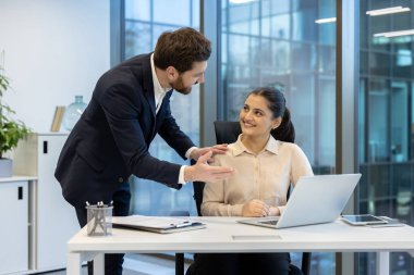 Bir iş kadını masasında oturmuş, dikkatle dinlerken meslektaşı parlak bir ofis ortamında rehberlik sunuyor..