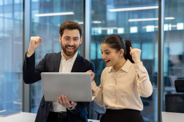Bir erkek ve kadın ofis ortamında dizüstü bilgisayara bakarken başarıyı kutluyorlar, mutlu ve heyecanlı. Modern binalar görülebilir..