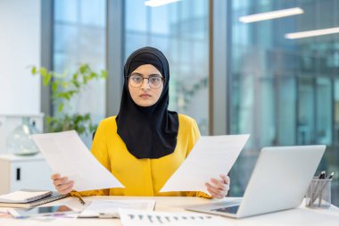 Başörtülü, ciddi görünümlü bir kadın bilgisayarını ve ofis ortamını kullanarak masasındaki belgeleri inceliyor..