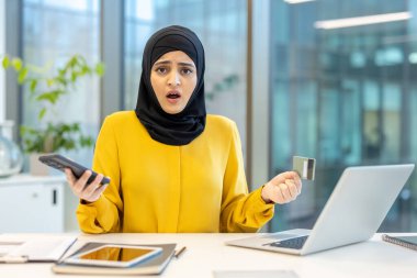Başörtüsü takan bir kadın, elinde bir kredi kartı ve telefon tutarken şaşırmış görünüyor. Ofiste bir masada oturuyor..