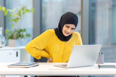Başörtüsü takan bir kadın, modern bir ofis ortamında dizüstü bilgisayarıyla çalışırken sırt ağrısı yaşar..
