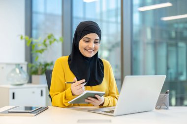 Başörtüsü takan gülümseyen bir kadın masasında çalışır, modern ofis ortamında dizüstü bilgisayar kullanırken notlar alır..