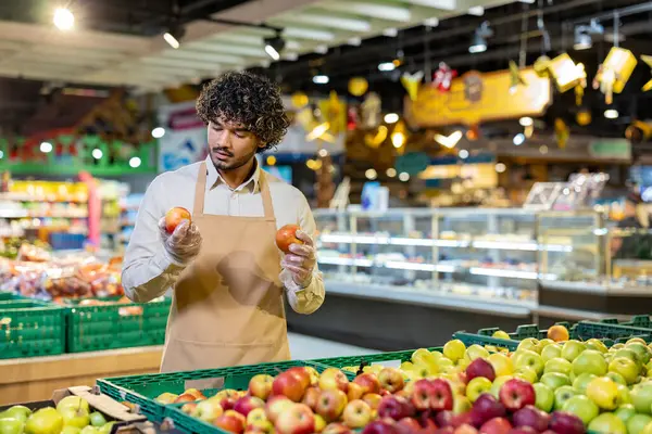 Bir market çalışanı üretim bölümünde bir çift elmayı dikkatle inceleyerek kalitelerini ölçer..