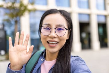 Asyalı bir kadın kameraya el sallarken gülümsüyor. Gözlük takıyor ve bir binanın dışında gibi görünüyor..