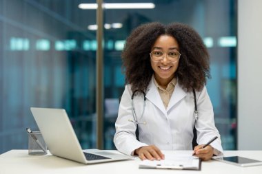 Masasında dostane ve gülümseyen siyah bir doktor çalışıyor. Gözlük ve steteskop takıyor..