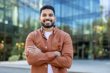 Gülümseyen bir adam kollarını kavuşturup modern cam bir binanın önünde poz verir. Açık hava, profesyonel ve ulaşılabilir bir ortam yakalıyor..