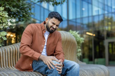 Bir adam bankta oturuyor, gözle görülür bir diz ağrısı yaşıyor, ifadesi rahatsızlık ve gerginlik gösteriyor. Fotoğraf gündüz vakti dışarıda çekiliyor..