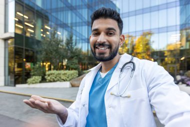 Neşeli bir doktor, modern bir cam binanın önünde seyirciyi davet eder ya da sunar..