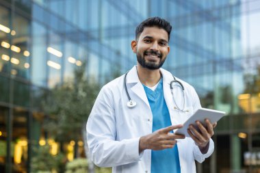 Hintli bir doktor modern bir cam binanın önünde tablet kullanırken gülümsüyor. Modern tıp ve teknolojiyi yansıtan bir sahne..