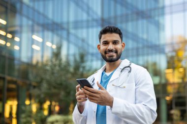 Gülümseyen bir doktor, modern bir ofis binasının yanında akıllı telefonunu açık havada tutarken kameraya bakar..