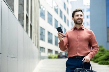 İyi giyimli bir adam bir elinde telefon, diğer elinde evrak çantası, gülümseyerek dışarı çıkar..