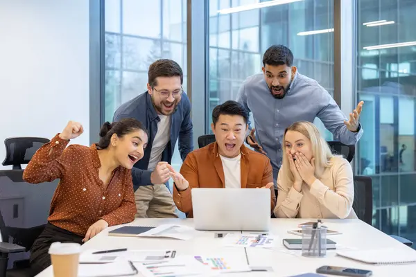 Çeşitli bir ekip başarılı bir projeyi heyecanlı bir şekilde dizüstü bilgisayar ekranına bakarak kutluyor. Takım çalışması ve başarı bariz.