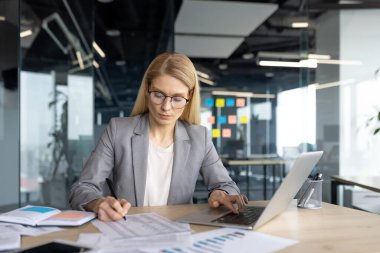 Modern ofis ortamında bir iş kadını, bir dizüstü bilgisayar üzerinde çalışırken ve masasındaki belgeleri incelerken, verileri analiz ederken,.
