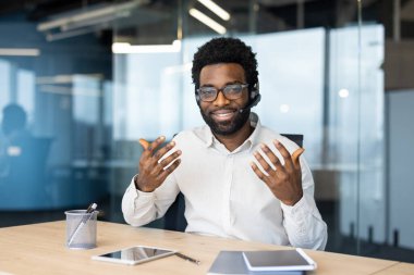 Genç bir adam, gözlük ve kulaklık takıyor, modern ofis ortamında bir video görüşmesi sırasında jestler yapıyor. Mutlu görünüyor..