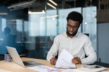 Modern bir ofiste masasında çalışan gözlüklü Afro-Amerikalı bir adam belgeleri inceliyor. Masada bir dizüstü bilgisayar ve bir tablet de var..