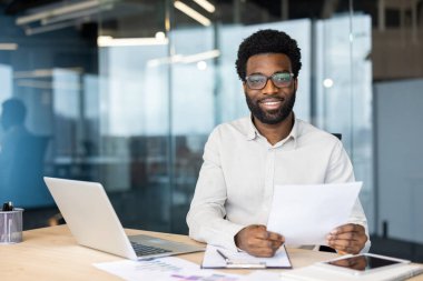 Ofis masasında çalışırken gülümseyen gözlüklü bir profesyonel belgeleri elinde tutuyor. Kameraya özgüven ve rahatlıkla bakar..