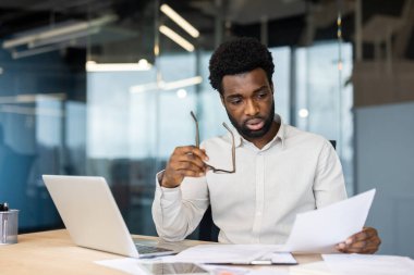 Odaklanmış siyah bir işadamı gözlüğünü tutarken, masasında özenle çalışırken belgeleri analiz eder. Bir ofiste oturuyor..