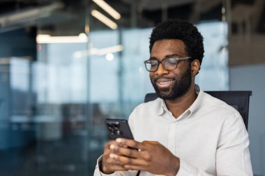 Gülümseyen gözlüklü bir adam modern bir ofiste otururken telefonuna bakar. İlginç bir şey okuyor ya da izliyor gibi görünüyor..