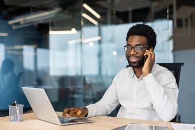 Afro-Amerikalı bir adam masasında çalışıyor, telefonuyla konuşuyor ve gülümserken bilgisayarında daktilo yazıyor. Ofis ortamında..