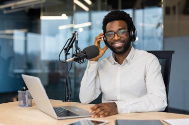 Podcast 'te gülümseyen, yakışıklı bir adam, gözlük ve kulaklık takıyor, kameraya bakıyor. Bir mikrofon, bir laptop ve bir masa var..