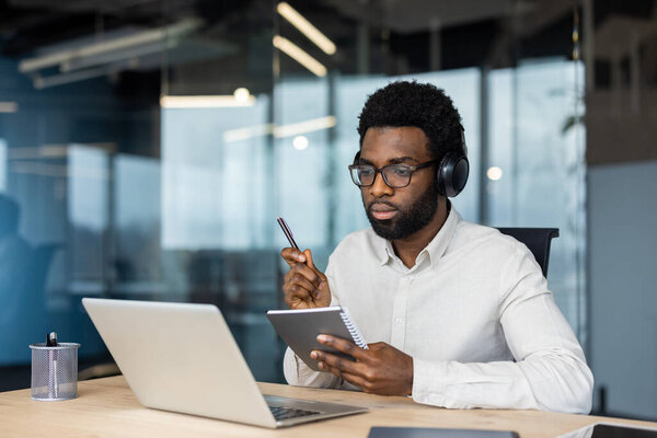 An african american man participates in a video conference while in his office, taking notes and working