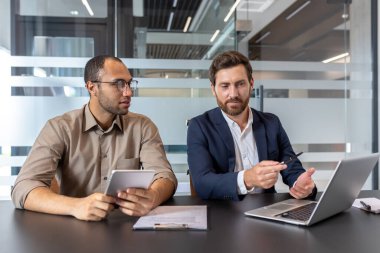 İki iş arkadaşı, belgeleri incelemek ve ofis masasında fikirleri tartışmak için bir dizüstü bilgisayar ve tablet kullanarak işbirliği yapıyorlar..