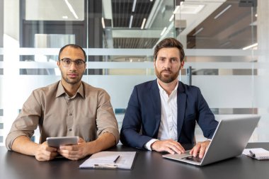 Kendine güvenen iki profesyonel masa başında oturuyor ve modern bir ofiste birlikte çalışmaya hazır görünüyorlar. Biri dizüstü bilgisayar kullanıyor..