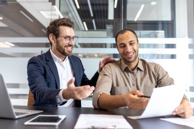 İki farklı iş adamı gülümsüyor, evrakları inceliyor ve modern ofis ortamında meseleleri tartışıyor. İşbirliği ve takım çalışması ortada..