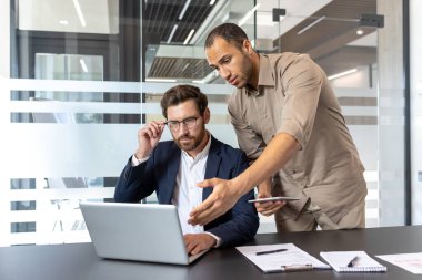 İki işadamı, bir dizüstü bilgisayarda sergilenen bir projeyi tartışmak için modern bir ofiste işbirliği yapıyorlar. Biri ekrana işaret ediyor.