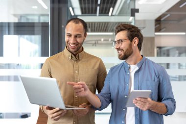 İki neşeli meslektaşı, modern ofis ortamında bir tabletin yardımıyla fikirleri tartışarak bir dizüstü bilgisayar üzerinde işbirliği yapıyorlar. Gülümsüyorlar..