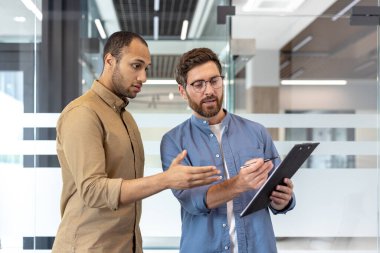 İki profesyonel evrak işlerini tartışıyor, biri işaret ediyor, diğeri de kalem tutuyor, modern bir ofis ortamında..
