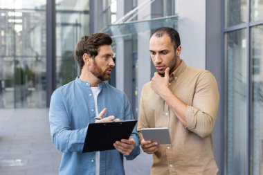 İki adam açık havada bir tartışma içindeler. Biri elinde bir pano tutarken, diğeri elinde bir tablet olduğunu düşünüyor..