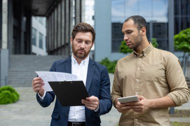 İki iş adamı belgeleri tartışıyor ve gün içinde modern bir ofis binasının dışındaki tablete bakıyorlar..