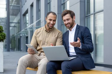 İki iş ortağı dışarıda oturuyor, bir dizüstü bilgisayar ve tablet üzerinde işbirliği yapıyor, işle ilgili verileri tartışıyorlar..
