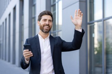 Bir iş adamı şehir merkezinde telefonunu tutarken el sallıyor. O, profesyonel kıyafetiyle mutlu ve başarılı görünüyor..