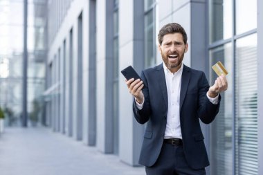 Hayal kırıklığına uğramış bir işadamı, elinde akıllı telefon ve kredi kartı olan bir ofis binasının önünde stresli bir durumda gibi görünüyor..