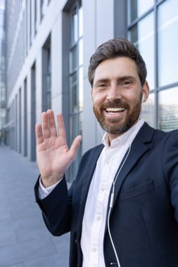 Takım elbiseli gülümseyen bir işadamı kulaklık takarken kameraya doğru el sallıyor. Arka planda bir ofis binası görünüyor..