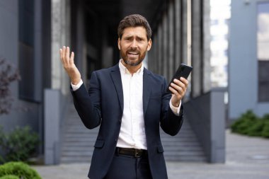 Takım elbiseli, hayal kırıklığına uğramış bir işadamı elinde telefon tutarken el kol hareketi yapıyor, muhtemelen dışarıda bir iş sorunu yaşıyor. Şehir binası arka planda..