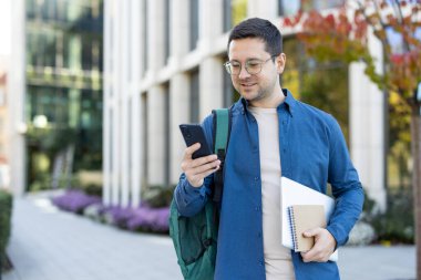 Genç bir adam dışarıda duruyor, akıllı telefonunu kontrol ediyor. Sırt çantası, defter ve dizüstü bilgisayar taşıyor, muhtemelen sınıfa giderken ya da çıkarken..