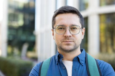 Gözlüklü ve sırt çantalı bir adamın portresi, kameraya tarafsız bir ifadeyle bakıyor..