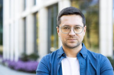 Modern bir binanın önünde gözlük takan bir adamın portresi. Mavi bir gömlek giyiyor, kameraya bakıyor..