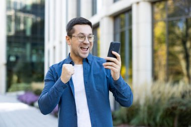 Dışarıda heyecanlı bir adam telefonuna bakıyor ve iyi haberi kutluyor. Gülümsüyor ve başarısını göstermek için sevinçle yumruğunu kaldırıyor..